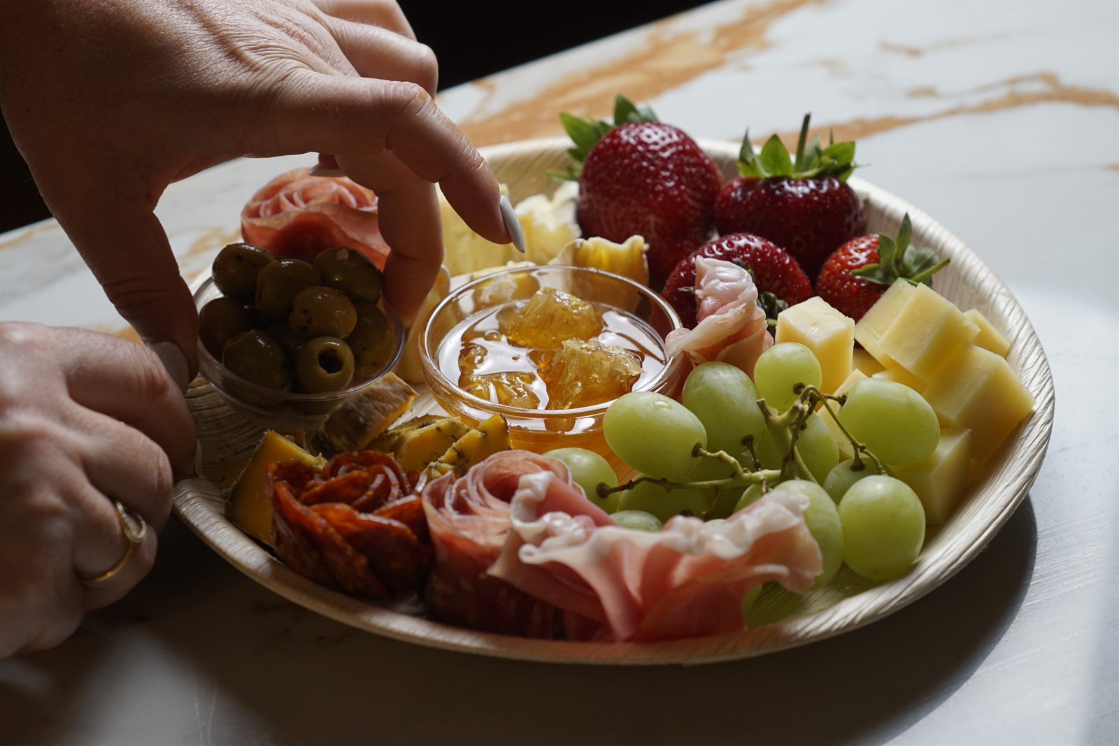 A guest reaching over a styled grazing board with honey, olives and charcuterie roses