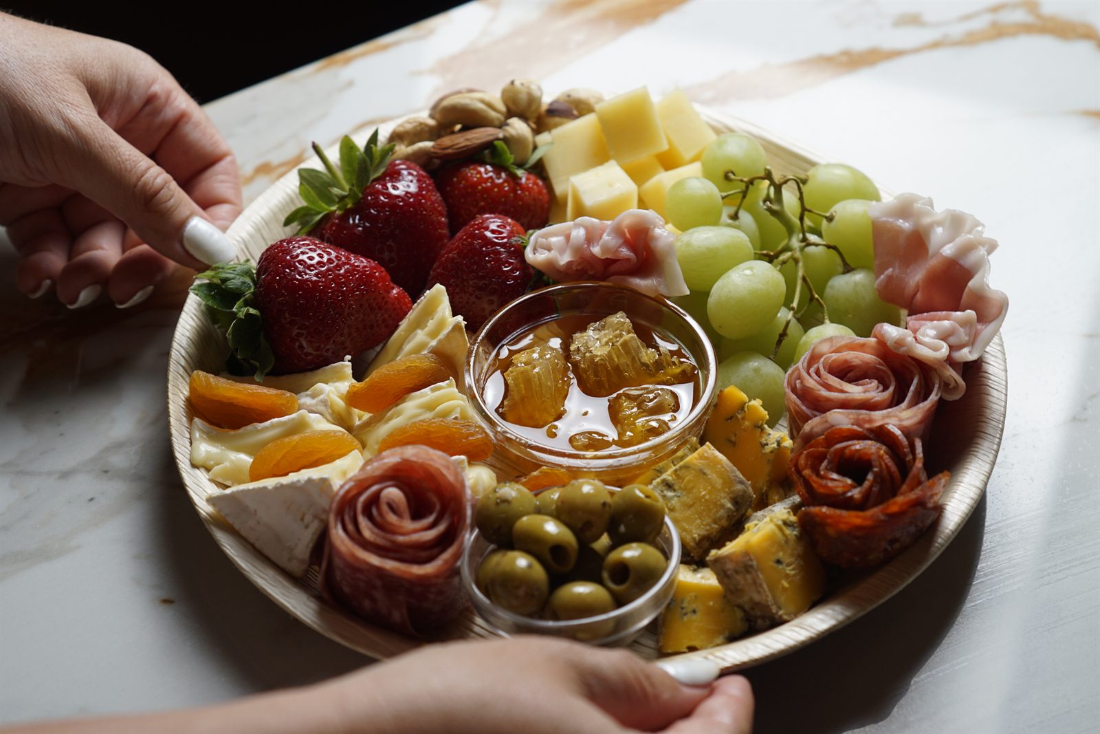 Two guests reaching for a shared grazing board with strawberries, honey and salami roses