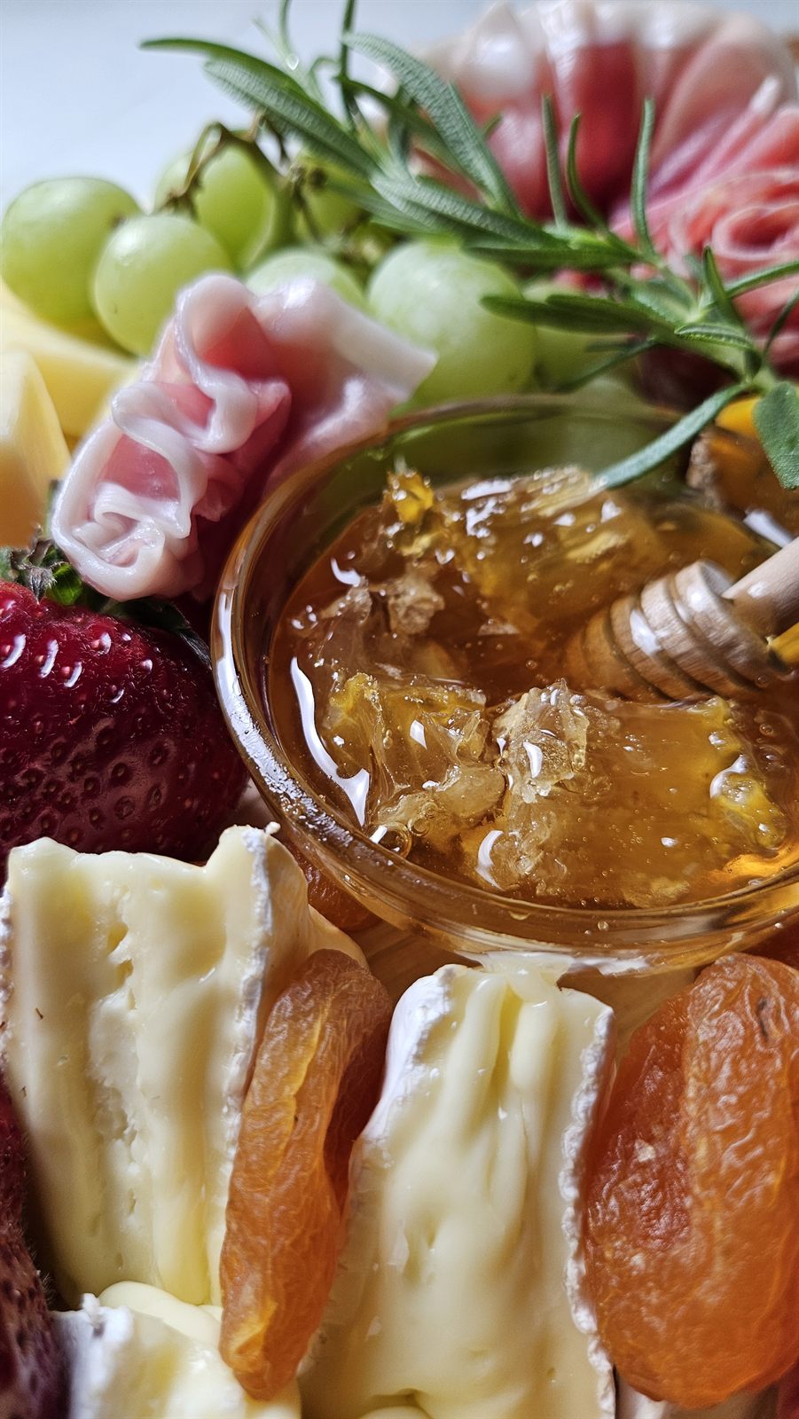A honeycomb dipper over a board with brie, dried apricots, prosciutto and grapes